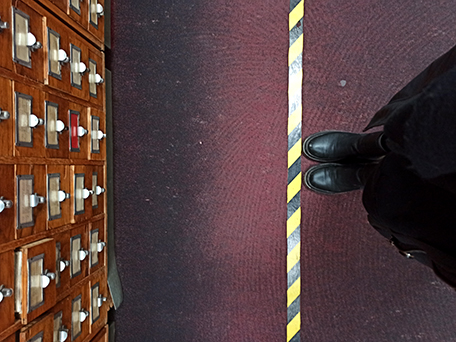 Blick von oben auf eine schwarz-gelbe Markierungslinie am Boden. Links steht ein alter hölzerner Karteikasten mit vielen Schubladen und weißen Knäufen, rechts sieht man die Füße einer Person in schwarzen Schuhen, die knapp an der Linie steht.