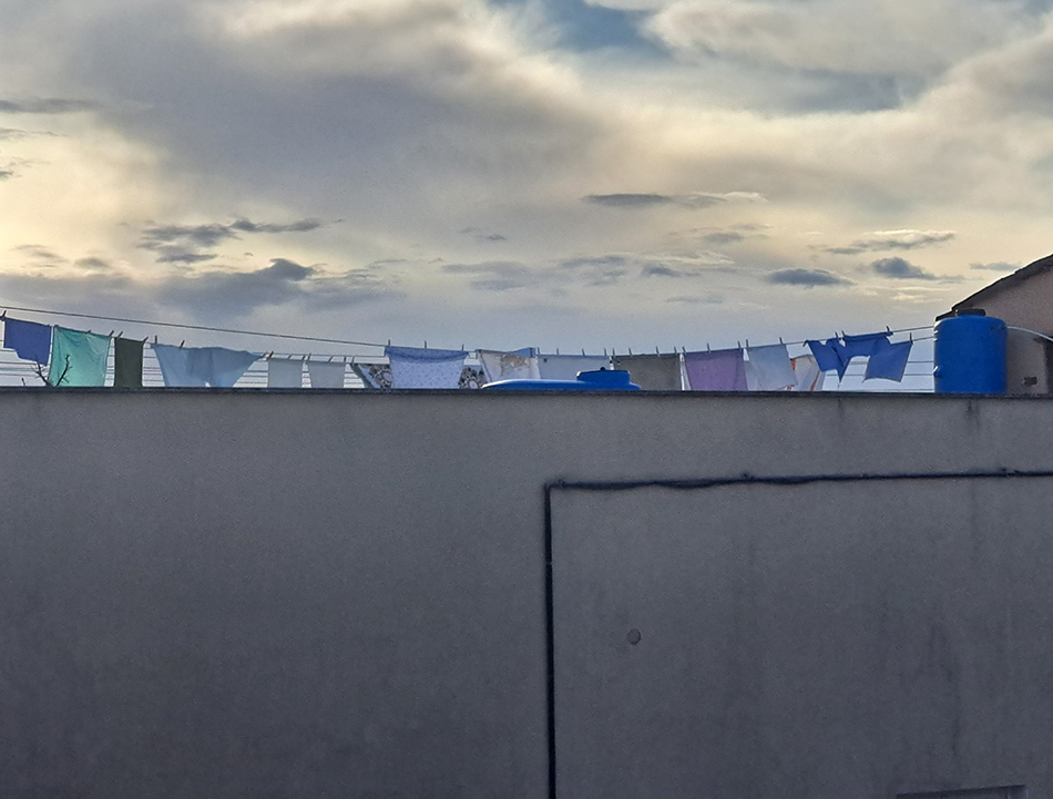 T-Shirts in Blautönen hängen an einer Wäscheleine und wehen im Wind, im Vordergrund ist eine große Mauer zu sehen, im Hintergrund leuchtet der Sonnenuntergang.