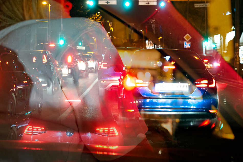 Aufnahme einer nächtlichen Verkehrsszene aus dem Auto heraus. Mehrere Fahrzeuge mit Rücklichtern stehen auf einer mehrspurigen Straße, Ampeln leuchten grün. Durch die Windschutzscheibe sind Spiegelungen von Lichtern und die Konturen einer Person sichtbar.