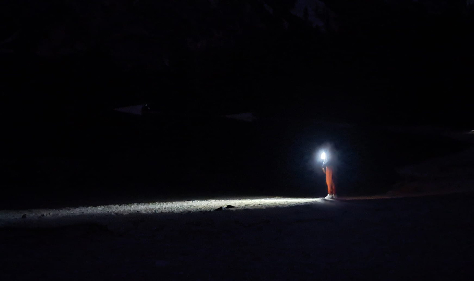 Eine Person steht in völliger Dunkelheit, nur die Stirnlampe auf ihrem Kopf beleuchtet die Szene und wirft einen schmalen Lichtkegel auf den Boden vor ihr.