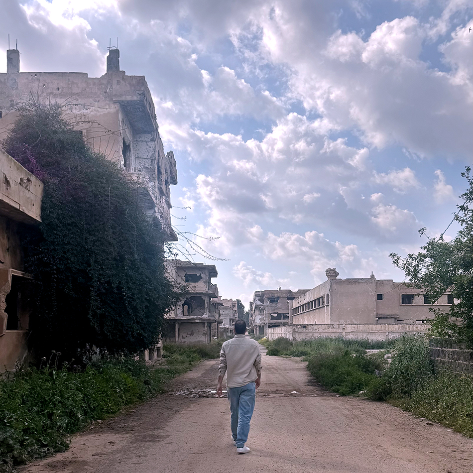 Eine überwucherte und zerbombte Straße in Damaskus, auf der ein Mann über den ungepflasterten Boden geht. An overgrown, bomb-damaged street in Damascus, with a man walking along the unpaved ground.
