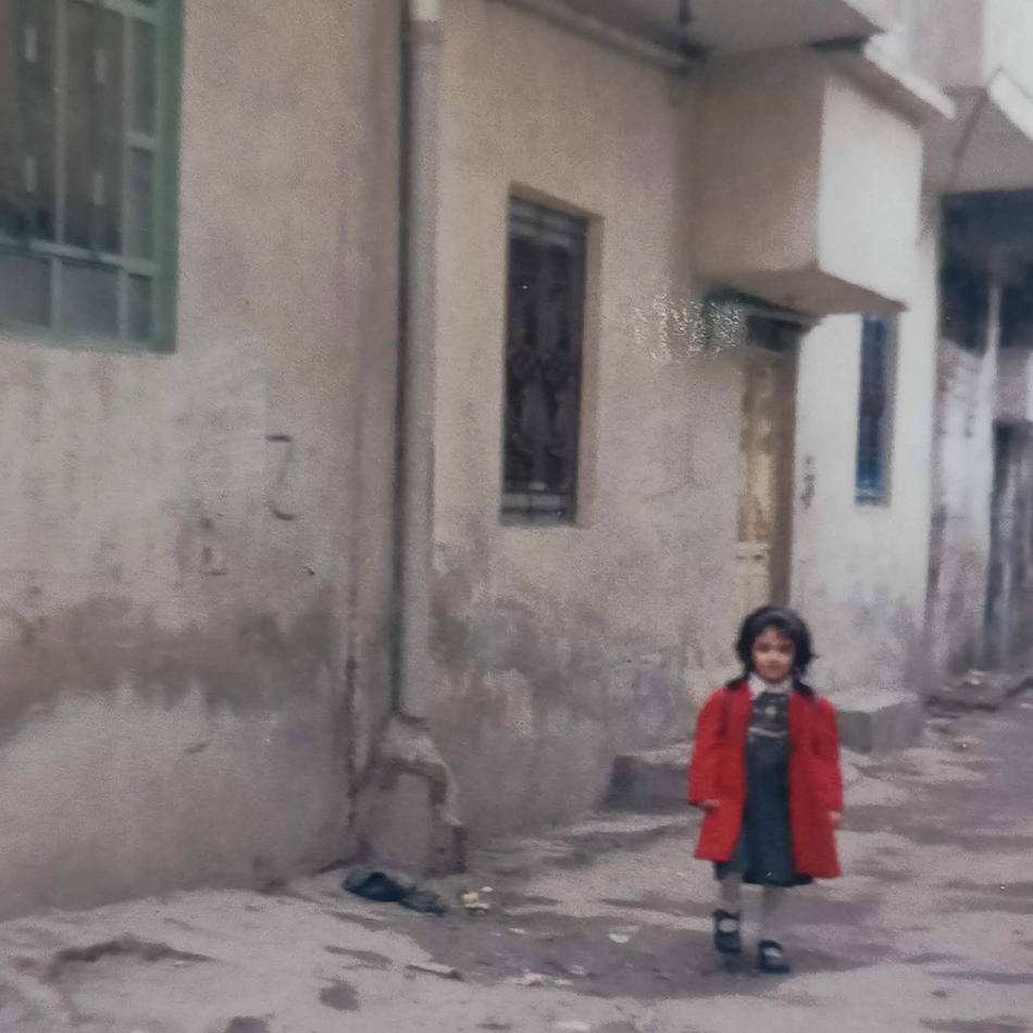 Analoge Farbfotografie eines etwa 3–4 Jahre alten Mädchens in einem roten Mantel, aufgenommen in den verwinkelten Gassen einer arabischen Stadt.Analog color photograph of a roughly 3–4-year-old girl in a red coat, taken in the narrow alleys of an Arab city.