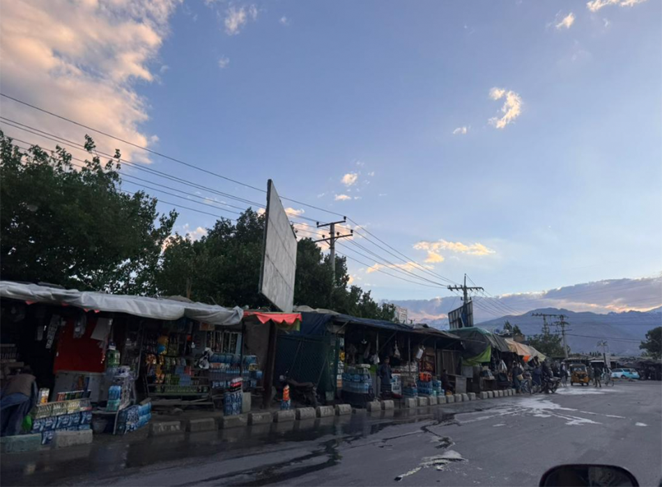 Straßenszene in Afghanistan bei Abenddämmerung, Verkaufsstände entlang einer nassen Straße, dahinter Berge. Street scene in Afghanistan at dusk, market stalls along a wet road with mountains behind.
