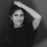 Schwarzweiß-Porträt von Shida Bazyar, eine Hand über dem Kopf, lächelnd in die Kamera. Black-and-white portrait of Shida Bazyar, one hand resting on her head, smiling at the camera.
