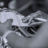 Symbolbild Schwarzweiß-Nahaufnahme zweier Hände, die einen Stift halten und in ein liniertes Heft schreiben. Im Hintergrund unscharf gemusterter Stoff. Symbolic image: Close-up of hands writing with a pen in a lined notebook.