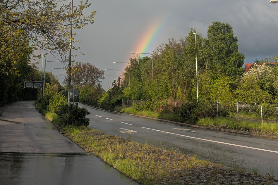 Ein Farbfoto von einer leeren Straße. Die ist vom Regen feucht. AmHimmel stehen dunkle Wolken, dazu erstreckt sich ein Regenbogen Richtung Himmel.