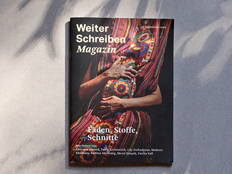 Das Weiter Schreiben Magazin liegt auf einer hellgrauen Fläche, über die Blattschatten fallen. Das Cover zeigt einen Oberkörper, eingehüllt in einen farbigen, gestreiften Stoff mit gestickten Ornamenten in Rot, Pink, Gelb und Grün. Zwei Hände halten den Stoff vor dem Körper. Titel: „Fäden, Stoffe, Schnitte